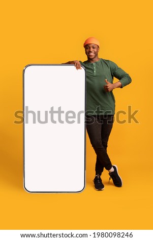 Happy Black Hipster Guy Leaning At Big Smartphone With Blank White Screen And Gesturing Thumb Up, Cheerful African Man Recommending New App Or Website, Standing On Yellow Background, Mockup Image