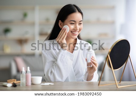 Cheerful asian woman cleaning her face, using cotton pads and cleansing product, looking at mirror in bedroom. Young attractive korean lady using face toner and cotton pad, home interior, empty space