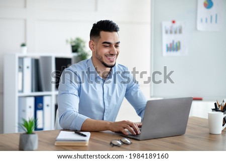 Handsome young Arab businessman working with laptop computer at his desk in office. Happy male entrepreneur typing on pc keyboard, checking email, taking part in online meeting at workplace