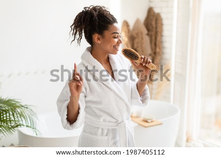 Happy Black Female Having Fun Singing With Hairbrush Brushing Hair And Enjoying Haircare Routine Standing In Modern Bathroom At Home, Wearing White Bathrobe.