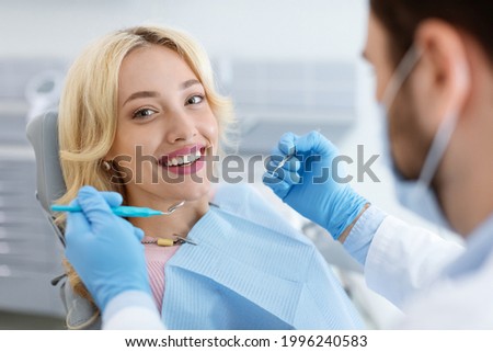 Closeup of man dentist in face mask doing treatment for patient cheerful young blonde lady smiling at camera, holding dental tools, wearing rubber gloves. Stomatology, modern dental clinic concept ストックフォト © 