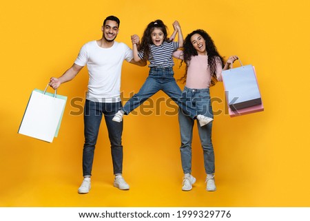 Portrait Of Excited Arab Parents And Little Daughter Holding Bright Shopping Bags And Having Fun Together Over Yellow Studio Background, Middle Eastern Family Enjoying Seasonal Shopping, Free Space 商業照片 © 