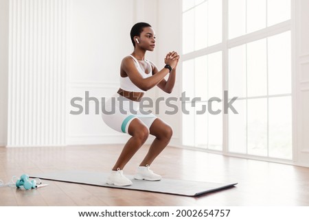 Body Shaping And Glutes Workout. Determined Fit Black Woman In White Sportswear Exercising With Resistance Loop Band Doing Squats Standing On Yoga Mat, Training Butt And Hamstring, Wearing Earbuds