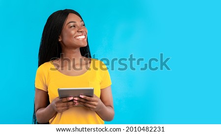 Happy black woman using digital tablet computer looking aside standing over blue studio background. Female browsing internet, looks at copy space. People and gadgets, e-learning. Panorama 商業照片 © 