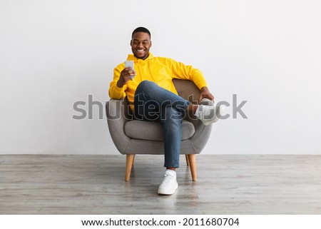 Great App. Contented Black Man Using Smartphone Advertising New Mobile Application, Texting Online Sitting In Armchair Near Gray Wall Indoors. Modern Mobile Communication, People And Gadgets