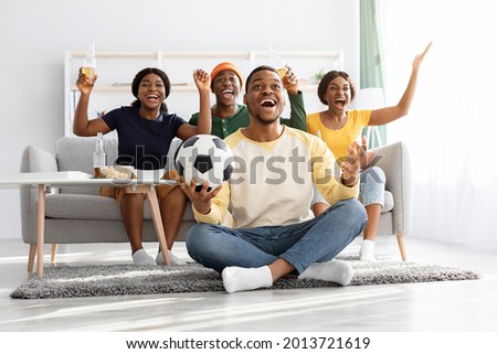Emotional football fans young african american men and women watching game together at home, celebrating win of their team, raising hands up and screaming, excited black guy with soccer ball