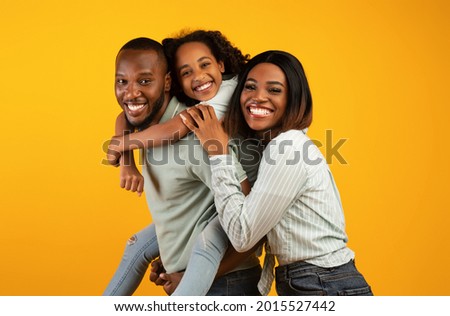Time with family. Overjoyed african american family laughing and posing to camera isolated over yellow studio wall. Cheerful father carrying his daughter on back 商業照片 © 
