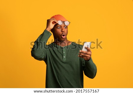 Amazing App. Shocked African American Guy Looking At Smartphone Screen, Amazed Black Millennial Man Taking Off Eyeglasses While Reading Unexpected Message, Standing Over Yellow Background, Copy Space
