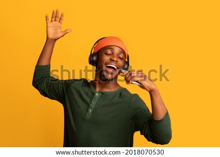 Technologies For Fun. Cheerful black hipster guy listening music in wireless headphones and singing, positive young african man using smartphone as microphone, having fun on yellow background