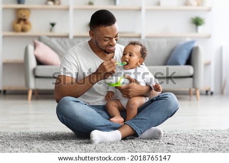 Caring Black Dad Feeding His Adorable Infant Baby From Spoon While Sitting Together On Carpet In Living Room, Young African American Father Enjoying Takic Care About Little Child, Copy Space Photo stock © 