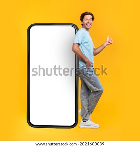 Excited Man Leaning On Big Smartphone With Blank White Screen And Gesturing Thumb Up Sign, Cheerful Guy Recommending New App Or Website, Standing On Yellow Background, Mock Up Image, Full Body Length