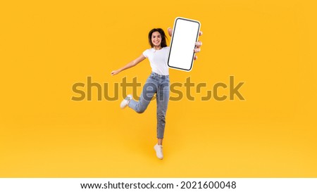 Happy young woman showing empty cellphone screen while jumping up on yellow studio background, collage, full body length, banner panorama. Excited lady recommending nice and useful mobile application