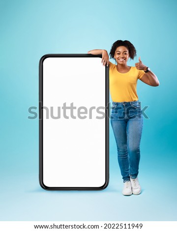 Mobile offer. Happy black lady leaning on huge cellphone with empty white screen, showing thumb up, recommending cool new app or website, offering space for your ad, mockup