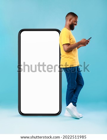 Full length of young Afro guy using cellphone while leaning on giant mobile phone with empty white screen, blue studio background. Mockup for app or website, space for advertisement