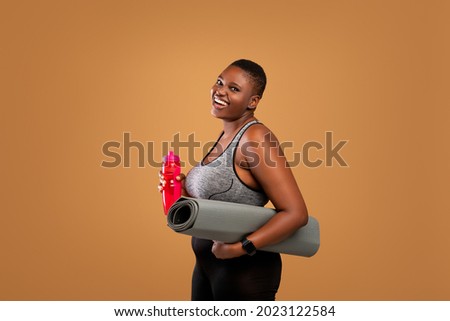 Fitness Concept. Portrait of happy black plus size woman holding water bottle and yoga mat, body positive female smiling and looking at camera standing isolated over brown studio background 商業照片 © 
