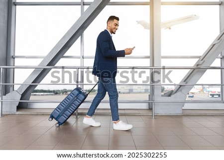Handsome arab guy using smartphone while walking with suitcase at airport terminal, young middle eastern man browsing mobile internet on cellphone while going to flight boarding, side view