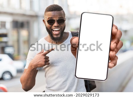 Great Offer. Portrait of excited black man standing outdoors holding big phone with white blank screen in hand, showing close to camera pointing at device. Gadget with empty free space for mock up