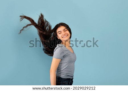 Portrait of smiling Indian teenage girl posing with long dark flying hair on blue studio background. Beautiful Eastern adolescent enjoying her healthy locks in blowing wind. Haircare concept