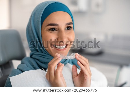 Orthodontics Concept. Smiling Muslim Woman In Hijab Holding Invisalign Or Invisible Braces While Sitting At Dentists Chair In Clinic, Islamic Female Using Clear Dental Aligner For Teeth Correction
