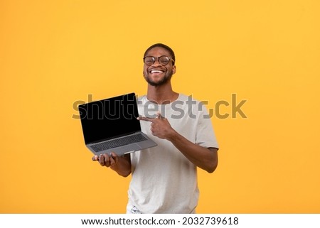 Joyful black man pointing at blank laptop screen with space for your website design over yellow studio background, mockup. African american guy showing PC with space for your advertisement 商業照片 © 