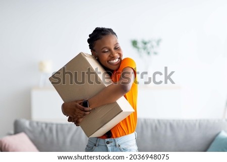 Cheerful Afro woman hugging carton parcel, receiving long awaited delivery, getting online order indoors. Satisfied female customer empracing her internet purchase in cardboard package