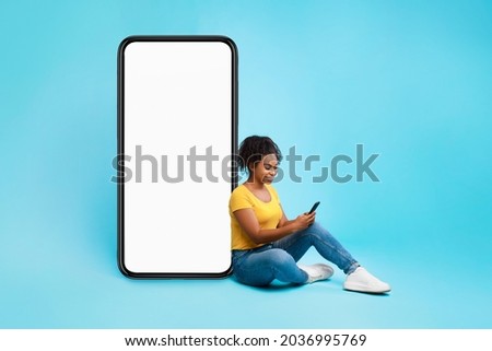 Cheery African American lady sitting near big cellphone with empty white screen, using mobile device, checking new cool app on blue studio background, mockup for website or application design