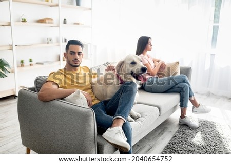 Upset young Arab man hugging pet dog after conflict with his girlfriend at home. Multiracial couple sitting on opposite sides of couch after quarrel, feeling unhappy and offended Foto stock © 