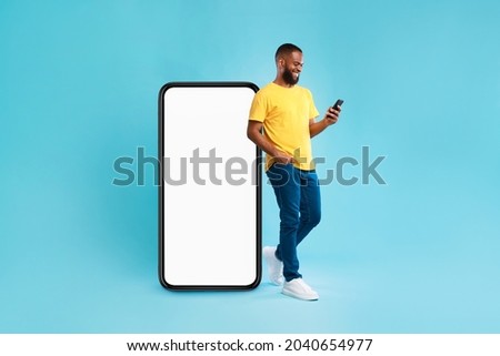 Handsome young Afro man using mobile phone while standing near huge smartphone with empty screen over blue studio background. Mockup for website or application, space for advertisement