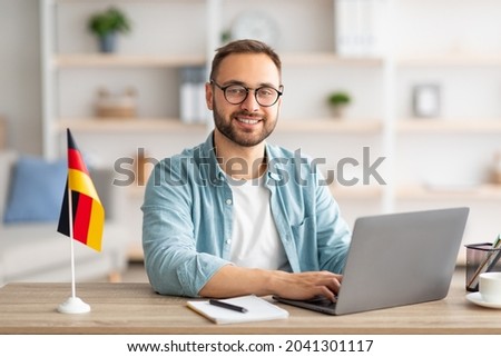 Happy young guy sitting at table with flag of Germany, using laptop pc, studying foreign language online at home. Modern remote education, abroad work and citizenship concept ストックフォト © 