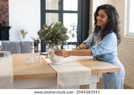 Modern home interior design, Scandinavian style concept. Beautiful young brunette woman arranging plants in vase, decorating dining room table, creating cozy atmosphere in her apartment, copy space