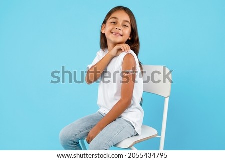 Vaccination Of Children. Happy Vaccinated Kid Girl Showing Arm With Adhesive Bandage After Vaccine Injection Sitting On Blue Studio Background. Kids And Covid-19 Prevention, Antiviral Immunization