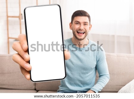 Mobile Offer. Joyful Young Man Showing Blank White Smartphone Screen At Camera While Sitting On Couch At Home, Cheerful Guy Showing Copy Space, Recommending New App Or Website, Mockup Image ストックフォト © 