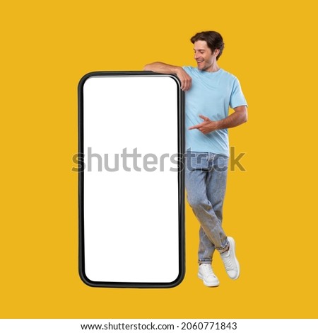 Mobile App Advertisement. Full Body Length Of Happy Man Leaning And Pointing At Big Huge White Empty Smartphone Screen Standing On Orange Studio Background. Check This Out, Cellphone Display Mock Up Stock foto © 