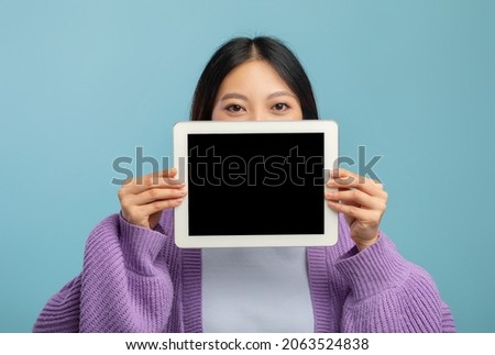 Young asian lady hiding her face behind digital tablet with empty black screen, showing cool mobile application or website on pad, mockup, blue studio background Imagine de stoc © 