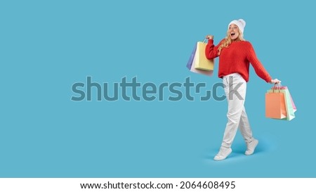 Seasonal Sales And Shopping. Full length of overjoyed female consumer walking and looking at free copy space, lady holding many colorful bags with new purchases isolated on blue studio wall, advert