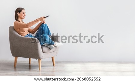 Young Caucasian lady with remote control sitting in armchair and watching TV against white wall, panorama with free space. Millennial woman with television controller switching television channels
