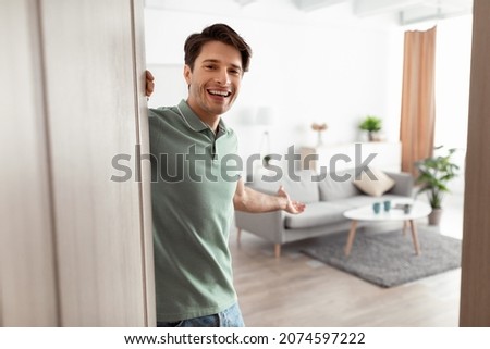 Welcome. Portrait of cheerful man inviting visitor to enter his home, happy young guy standing in doorway of modern apartment, millennial male holding door looking out showing living room with hand Foto stock © 