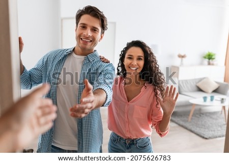 Portrait of cheerful couple inviting guests to enter home, happy young guy and lady standing in doorway of modern flat, looking out, man shaking hands, meeting new neighbors or friends
