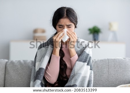 Unhappy sad young indian female in plaid suffering from fever and flu on sofa, blowing nose in napkin in living room interior. Covid-19 lockdown, treatment of illness, cold and runny, copy space Сток-фото © 