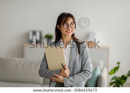 Portrait of female arab psychotherapist with clipboard looking aside and smiling, standing at modern office, free space. Professional psychoanalyst providing psychological services at clinic Foto d'archivio © 