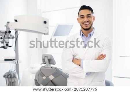 Dental Center. Portrait Of Smiling Middle Eastern Dentist Doctor Posing At Workplace, Handsome Arab Stomatologist Standing With Folded Arms In Modern Clinic Interior, Ready For Check Up With Patient Foto stock © 
