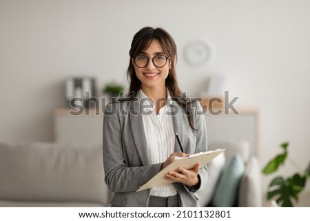 Happy professional middle eastern female psychologist writing in clipboard, looking and smiling at camera, working in modern office. Psychotherapy services, mental health professional concept Stockfoto © 