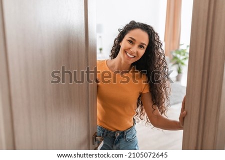 Portrait of cheerful young lady standing in doorway of new modern home, receiving and greeting visitor, happy smiling curly lady holding door looking out of slightly open ajar door Foto stock © 