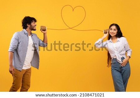 Photo of Love message. Loving indian man telling romantic words to girlfriend through tin can phone with heart shaped string, standing over yellow background, creative collage