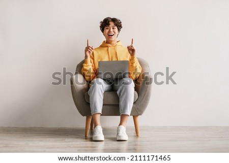 Millennial Asian man having creative idea, gesturing eureka with both hands, pointing up, offering space for design, sitting in armchair with laptop pc against white studio wall, copy space Сток-фото © 