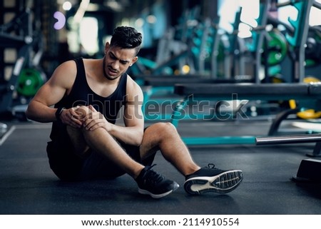 Sport Trauma. Young Arab Male Athlete Touching His Hurted Knee At Gym, Upset Middle Eastern Guy Sitting On Floor And Massaging Sore Injured Leg After Workout Training In Fitness Club, Free Space Stock foto © 