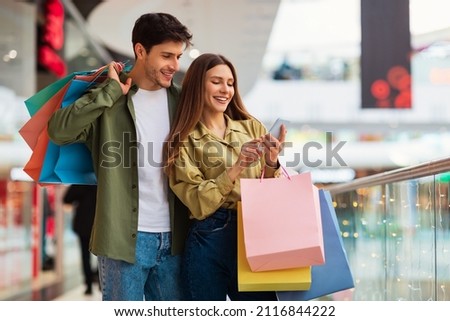 Buyers Couple Shopping Using Cellphone Holding Colorful Shopper Bags Standing In Mall. Happy Customers Using Application Purchasing Clothes Online Via Smartphone. Ecommerce And Shopaholism Imagine de stoc © 
