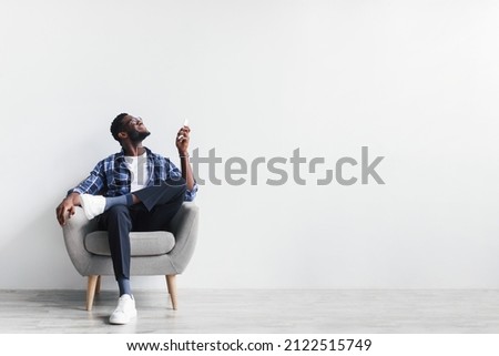 Split system. Cheerful young black man with remote turning on air conditioner, sitting in armchair against white studio wall, mockup. Happy millennial African American guy using climate control 商業照片 © 