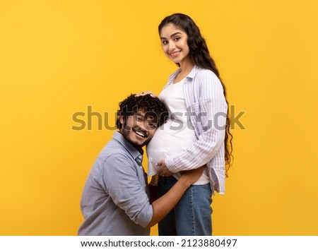 Caring indian man putting his ear on wife's pregnant tummy, listening to baby's heartbeat, yellow studio background. Millennial family expecting child, expressing love to each other Foto d'archivio © 