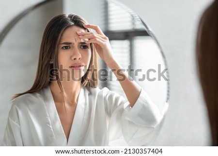 Unhappy Young Woman Looking In Mirror And Touching Wrinkles On Her Face, Attractive Millennial Female Standing In Bathroom And Examining Skin Fine Lines, Selective Focus On Reflection, Closeup Foto stock © 